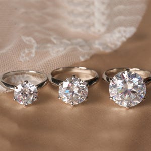 May include: Three silver rings with large, clear, round-cut gemstones. The rings are set on a neutral-toned surface, with a delicate lace fabric in the background. The gemstones sparkle, reflecting light.
