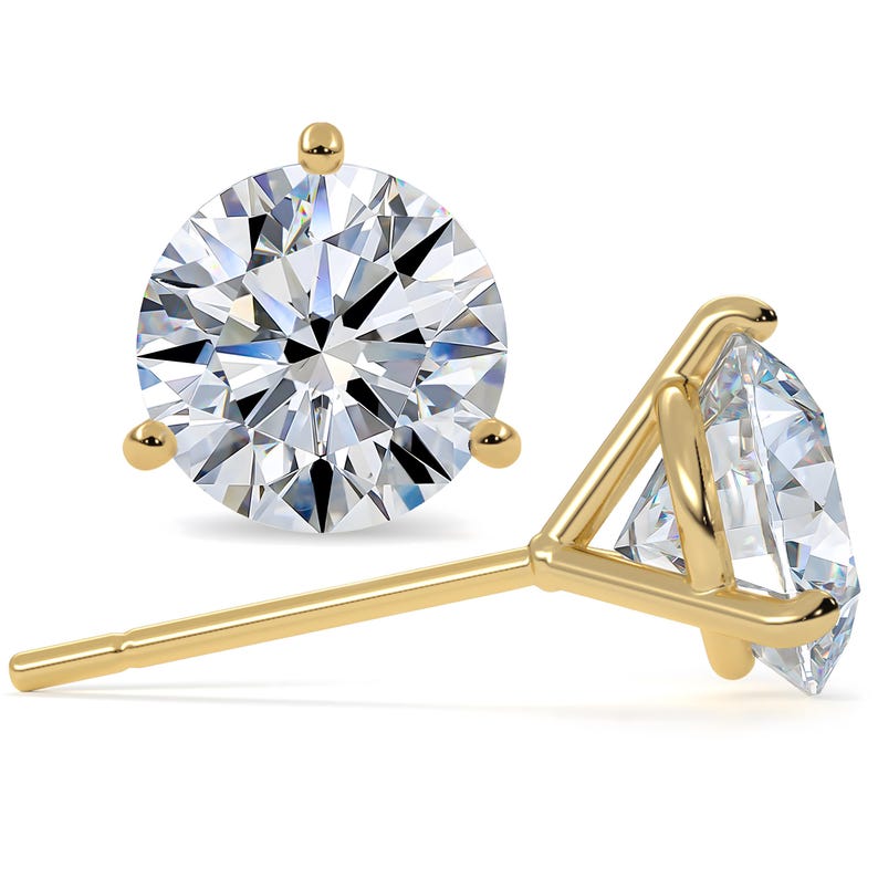 May include: Close-up of two gold stud earrings with a three-prong setting. Each earring features a round, brilliant-cut diamond. The gold setting and diamond facets reflect light, creating a sparkling effect. The earrings are set against a white background.