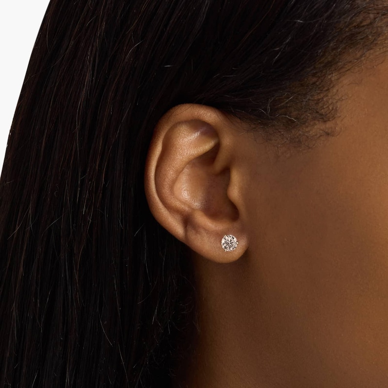 May include: A close-up of a round, sparkling diamond stud earring. The earring is set in a simple, classic design. The earring is shown on a person's ear, with dark hair in the background.