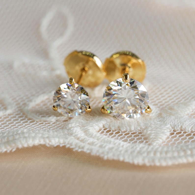 May include: A pair of gold stud earrings with large, clear, round-cut gemstones. The earrings are set in a gold prong setting and are displayed on a white lace surface. The earring backs are also gold.