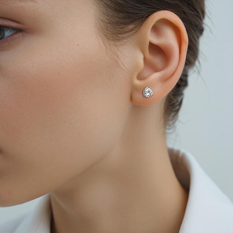 May include: A close-up of a person's ear with a single, round, clear gemstone stud earring. The earring is set in a silver or white metal setting. The earring is the focal point of the image.