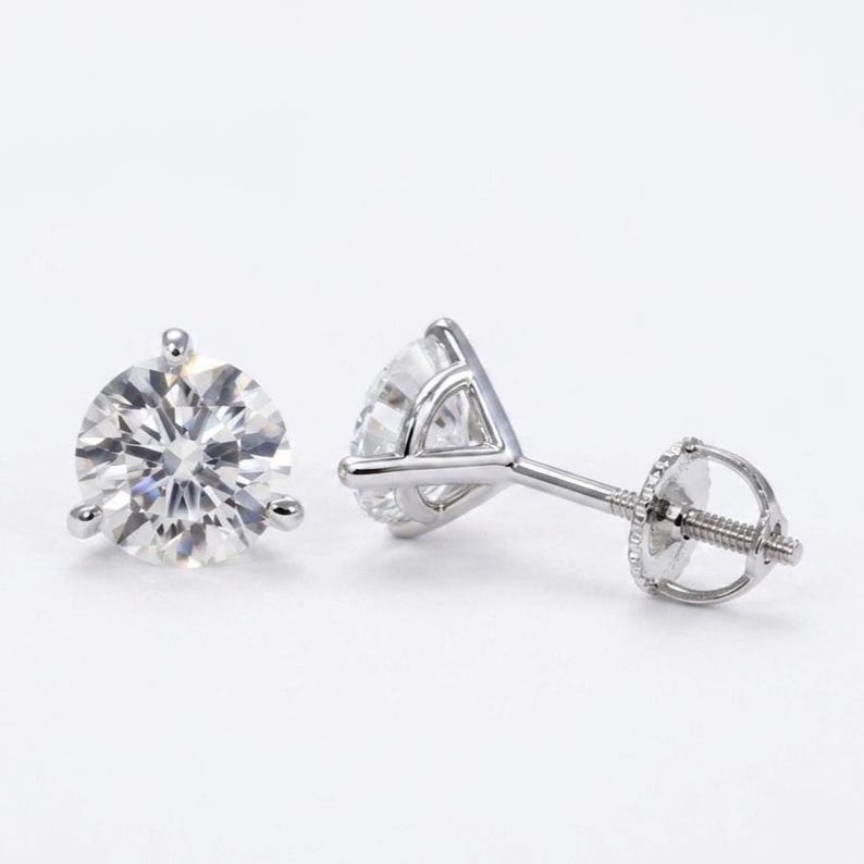 May include: A pair of sparkling round-cut diamond stud earrings. Each earring features a brilliant, clear gemstone set in a silver-colored, three-prong setting with a screw-back closure. The earrings are displayed against a white background.