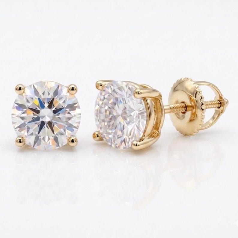 May include: A pair of round-cut diamond stud earrings with a gold setting. The earrings feature a brilliant, clear gemstone held in place by four prongs. The post and backing are also gold.