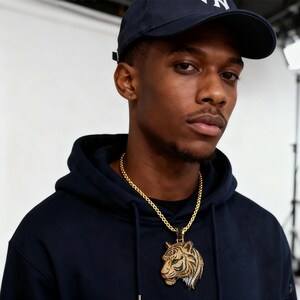 May include: A gold chain necklace with a tiger pendant. The pendant features a detailed tiger head with green eyes, black and white accents, and a gold frame. The person in the image is wearing a navy blue hoodie and a navy blue baseball cap.