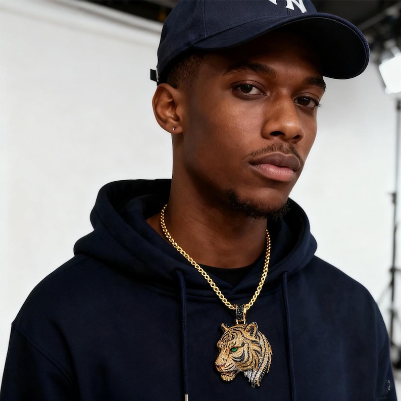 May include: A gold chain necklace with a tiger pendant. The pendant features a detailed tiger head with green eyes, black and white accents, and a gold frame. The person in the image is wearing a navy blue hoodie and a navy blue baseball cap.
