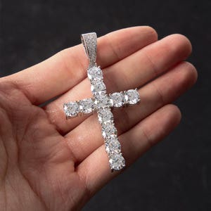 May include: A silver-toned cross pendant featuring a series of round, clear, faceted stones set along the entire length of the cross arms. The top bail is also encrusted with smaller, clear, sparkling stones, creating a bright, reflective finish.