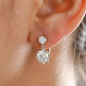 May include: A close-up of a silver earring with two clear, round-cut gemstones. The top gemstone is smaller, and the bottom gemstone is larger and hangs below it. The earring is attached to the ear with a silver hook.