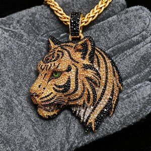 May include: A gold-colored pendant necklace featuring a tiger head design. The tiger's face is adorned with black and gold-colored gemstones, with a green gemstone for the eye. The necklace chain is also gold-colored.