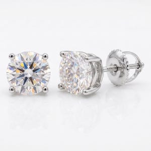 May include: A pair of round-cut diamond stud earrings. The earrings feature a brilliant, clear gemstone set in a four-prong setting. The posts and backs are made of a shiny, silver-colored metal. The earrings are displayed on a white background.