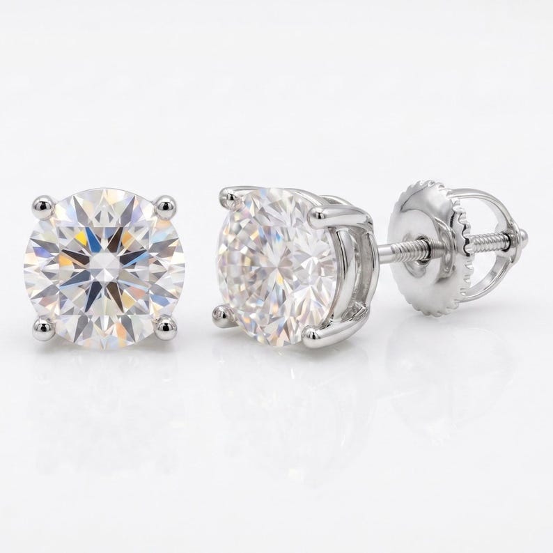 May include: A pair of round-cut diamond stud earrings. The earrings feature a brilliant, clear gemstone set in a four-prong setting. The posts and backs are made of a shiny, silver-colored metal. The earrings are displayed on a white background.