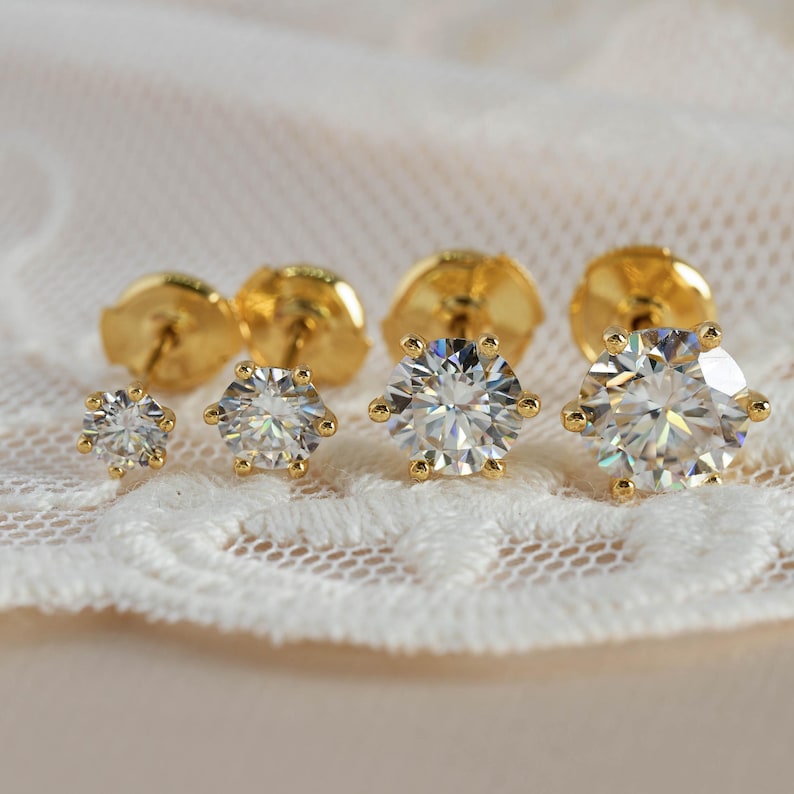 May include: Close-up of three pairs of gold stud earrings with clear, faceted gemstones. The earrings are set in gold with a six-prong setting. The earrings are displayed on a white lace surface.