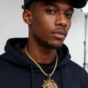 May include: A gold chain necklace with a tiger pendant. The pendant features a detailed tiger head with green eyes, black stripes, and a gold and silver finish. The person in the image is wearing a navy blue hoodie and a navy blue baseball cap.