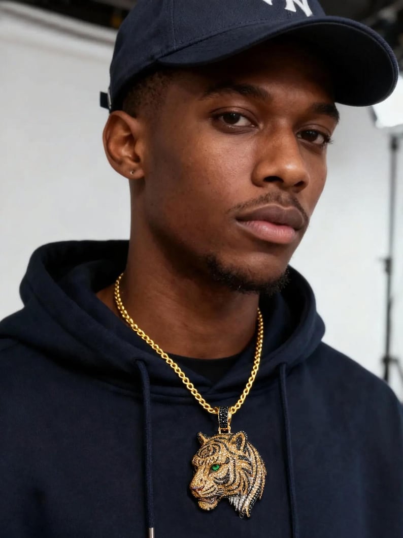 May include: A gold chain necklace with a tiger pendant. The pendant features a detailed tiger head with green eyes, black stripes, and a gold and silver finish. The person in the image is wearing a navy blue hoodie and a navy blue baseball cap.