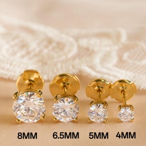 May include: Four pairs of gold-colored stud earrings with clear, round gemstones. The earrings are displayed in a row, with sizes 8mm, 6.5mm, 5mm, and 4mm labeled below. The background is a soft, neutral color.