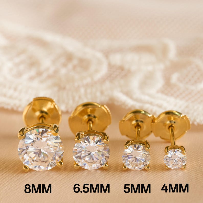 May include: Four pairs of gold-colored stud earrings with clear, round gemstones. The earrings are displayed in a row, with sizes 8mm, 6.5mm, 5mm, and 4mm labeled below. The background is a soft, neutral color.