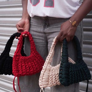 May include: Four handmade crochet bags in black, red, cream, and dark green. The bags have a rounded shape with a thick, braided handle. The bags are held by a person wearing a white shirt with the number "07" on it.