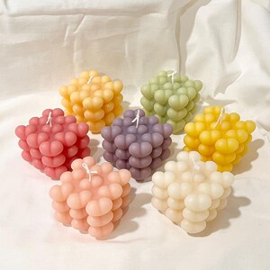 May include: Six colorful bubble-shaped candles in various pastel colors, including pink, yellow, green, purple, and cream. The candles are arranged in a circle on a white surface.