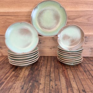 May include: A collection of ceramic plates with a scalloped edge. The plates have a light green and brown color scheme. Several stacks of plates are visible, along with a few individual plates. The plates are arranged on a wooden surface.