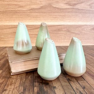 May include: Four ceramic salt and pepper shakers. The shakers are cone-shaped with a light green glaze and a brown base. They are arranged on a wooden surface with a vintage book.