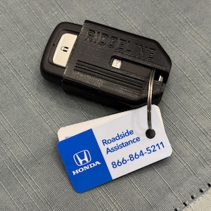 May include: Black key fob cover with "RIDGELINE" text, holding a car key. A white and blue tag with "Roadside Assistance" and a phone number is attached to the key ring. The background is a grey textured surface.