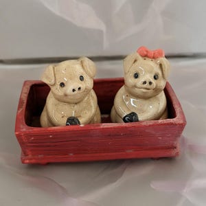 May include: A pair of ceramic pig figurines in a red wagon. The pigs are beige with black eyes and hooves. One pig has a pink bow. The wagon is rectangular and painted red.