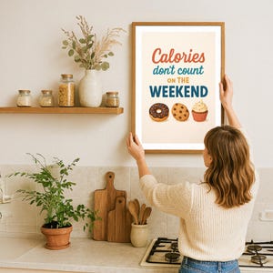 Op de afbeelding: Een ingelijst print met de tekst "Calories don't count on the weekend" in blauwe en rode letters. De print toont een donut, een koekje en een cupcake.