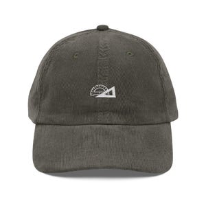 May include: A dark olive green corduroy baseball cap with a curved brim. The cap features a small white embroidered design of a protractor and triangle on the front. The hat has a structured fit and a top button.