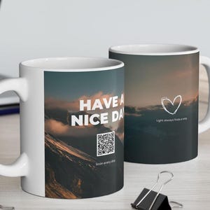 May include: Two white ceramic mugs with a mountain landscape design. One mug features the text "HAVE A NICE DAY" with a QR code and "Scan every day." The other mug has a heart and the text "Light always finds a way."