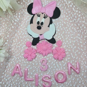 May include: Pink fondant cake topper with a cartoon Minnie Mouse, flowers, and the name "Alison" with the number "3" in pink frosting.