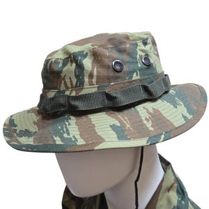 May include: Camouflage-patterned boonie hat in shades of green, brown, and tan. The hat features a wide brim, a fabric band around the crown, and two ventilation eyelets. A chin strap is attached. This hat is designed for outdoor activities.