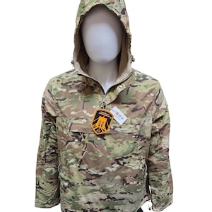May include: Camouflage anorak jacket with a hood and a half-zip closure. The jacket features a multi-tone pattern of greens, browns, and tans. A tag with text and a logo is attached to the zipper. The jacket is displayed on a mannequin.
