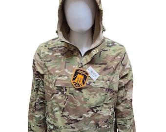 May include: Camouflage anorak jacket with a hood and a half-zip closure. The jacket features a multi-tone pattern of greens, browns, and tans. A tag with text and a logo is attached to the zipper. The jacket is displayed on a mannequin.