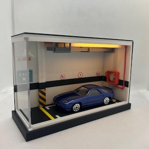 May include: A blue toy car parked in a miniature garage diorama with a white ceiling, black and white striped floor, and a yellow exit sign that reads "EXIT".