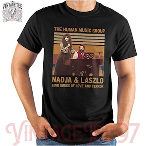 May include: Black t-shirt featuring a vintage-style graphic of Nadja and Laszlo from "The Human Music Group." The design includes text that reads "Sing Songs of Love and Terror." The shirt has a classic crew neck and short sleeves.