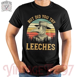 Puede incluir: Camiseta negra con un diseño de médico de la peste con máscara y sombrero, sobre un fondo de puesta de sol. El texto dice "BUT DID YOU TRY LEECHES". El logotipo de Vintage Tee 97 es visible.