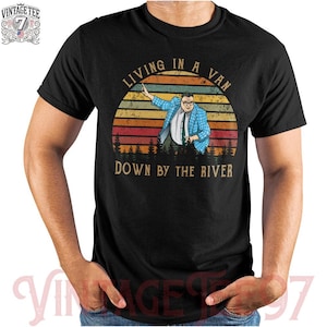 May include: Black t-shirt with a graphic of a man in a blue plaid jacket, glasses, and a tie, set against a sunset backdrop with the text "LIVING IN A VAN DOWN BY THE RIVER". The shirt has a vintage logo.