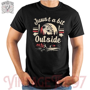 Puede incluir: Camiseta negra con un gráfico de béisbol vintage y el texto "Just a bit Outside".