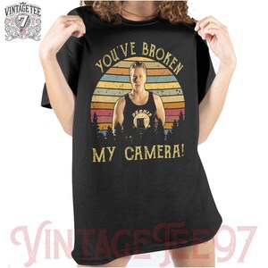 May include: A black t-shirt with a vintage-style graphic of a man with a camera. The graphic features a sunset background with the text "You've Broken My Camera!" in white.