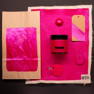 Pode incluir: Uma amostra de tecido rosa com um pequeno frasco de tinta rosa etiquetado como "toune" e um saco de papel castanho com uma amostra de tecido rosa dentro. A amostra de tecido tem um pequeno círculo rosa, uma etiqueta castanha com uma faixa rosa e um círculo castanho com um furo no centro. A amostra de tecido está etiquetada como "#311" e o saco de papel está etiquetado como "SM 311".