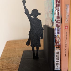 May include: A black metal bookend featuring a silhouette of a character wearing a straw hat with one arm raised in the air. The bookend has a flat rectangular base and a vertical support panel, currently holding a One Piece manga volume.