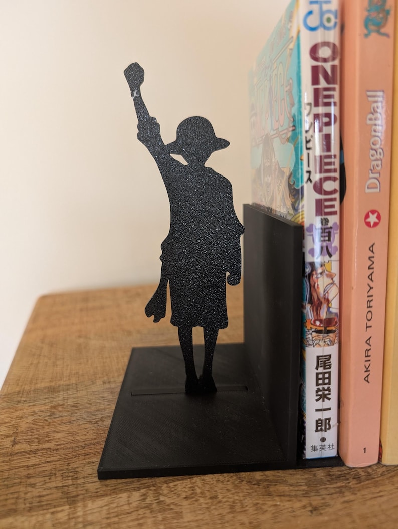 May include: A black metal bookend featuring a silhouette of a character wearing a straw hat with one arm raised in the air. The bookend has a flat rectangular base and a vertical support panel, currently holding a One Piece manga volume.