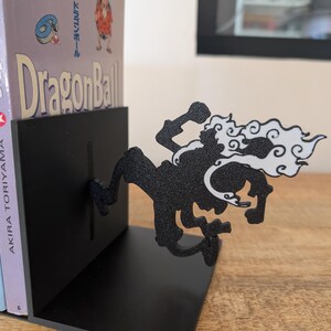 May include: A black metal bookend featuring a silhouette of a character in a dynamic pose with white cloud-like accents. The bookend supports a copy of the Dragon Ball manga by Akira Toriyama, which shows the title and a small character illustration.
