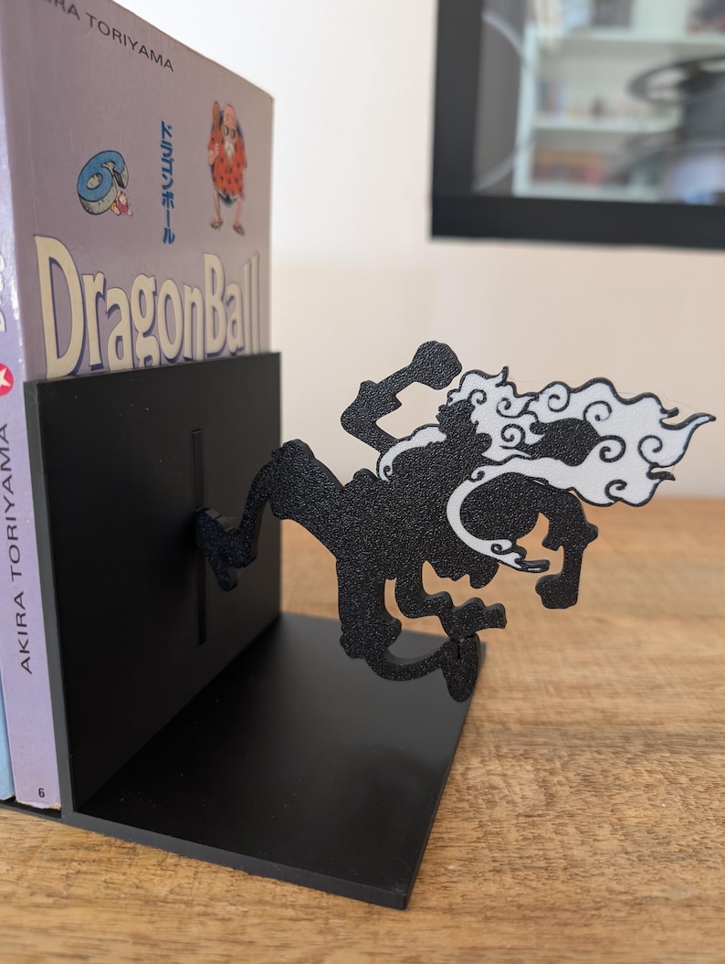 May include: A black metal bookend featuring a silhouette of a character in a dynamic pose with white cloud-like accents. The bookend supports a copy of the Dragon Ball manga by Akira Toriyama, which shows the title and a small character illustration.