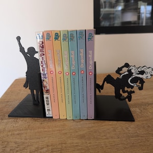 May include: A set of two black metal bookends featuring silhouettes of anime characters, holding a row of seven manga volumes. The books include one One Piece volume and six Dragon Ball volumes by Akira Toriyama, arranged in a colorful sequence.