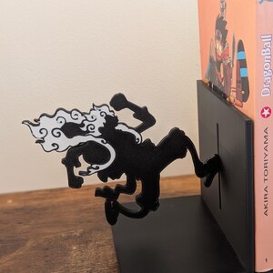 May include: A black metal bookend featuring a silhouette of a running character with white hair accents. The bookend has a flat base and a vertical plate that supports a copy of Dragon Ball by Akira Toriyama, which shows a character in a helicopter.