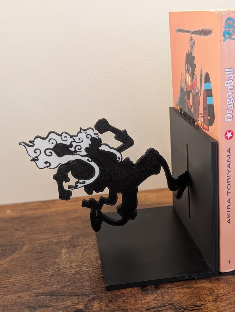 May include: A black metal bookend featuring a silhouette of a running character with white hair accents. The bookend has a flat base and a vertical plate that supports a copy of Dragon Ball by Akira Toriyama, which shows a character in a helicopter.