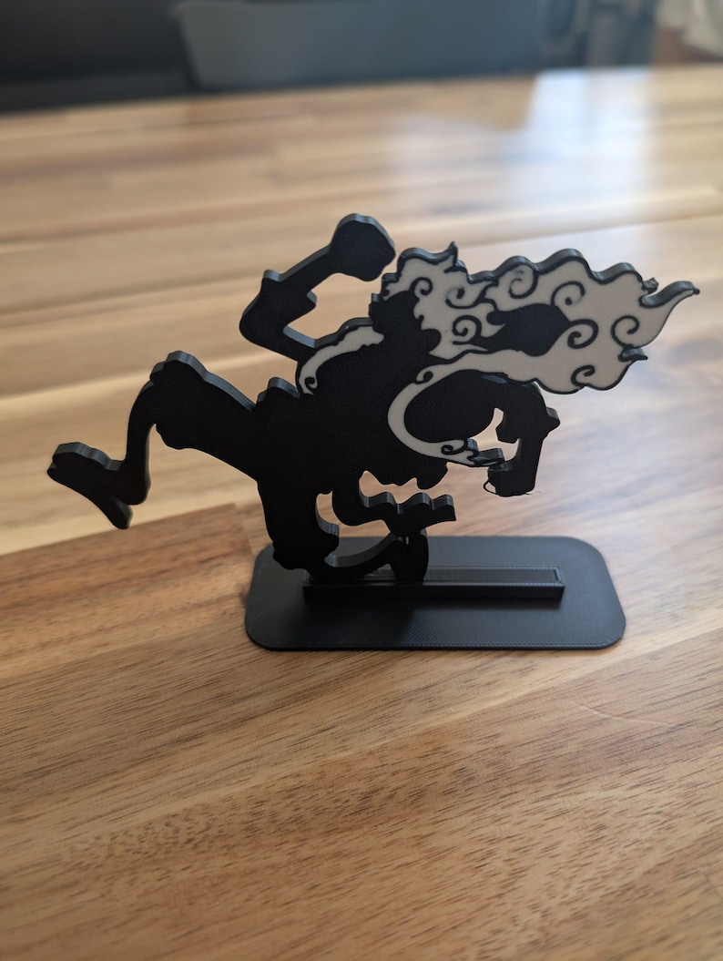 May include: A black 3D printed figurine of a dynamic character in a running pose, featuring a white cloud-like accent trailing from the head and shoulders. The figure is mounted on a flat, rectangular black base for stability and display.