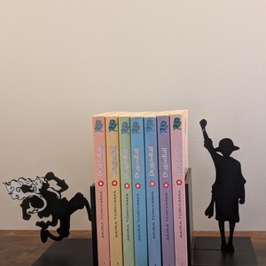 May include: A set of black metal bookends featuring silhouettes of anime characters, one in a dynamic leaping pose and the other standing with a raised fist, holding seven Dragon Ball manga volumes by Akira Toriyama with pastel-colored spines.