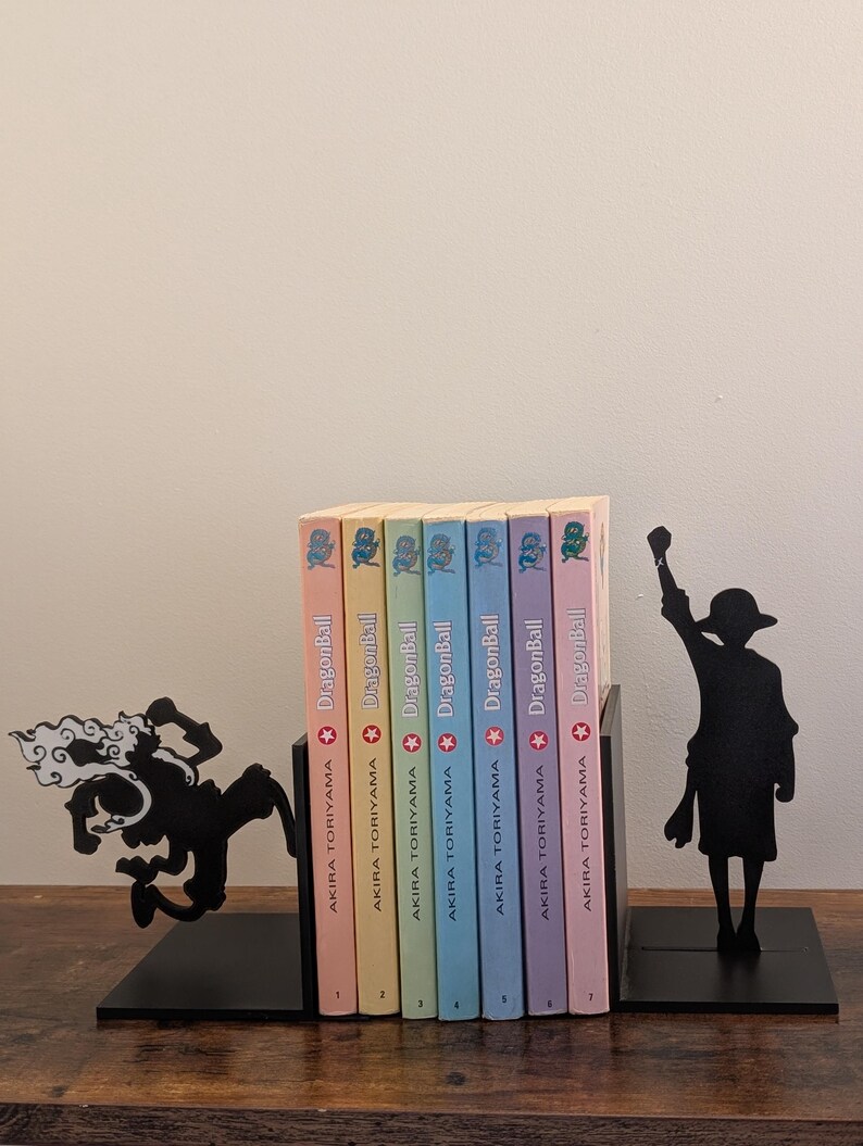 May include: A set of black metal bookends featuring silhouettes of anime characters, one in a dynamic leaping pose and the other standing with a raised fist, holding seven Dragon Ball manga volumes by Akira Toriyama with pastel-colored spines.