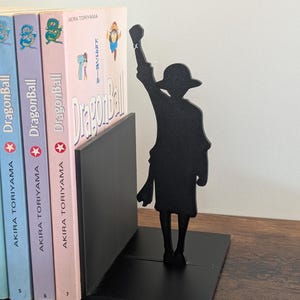 May include: A black metal bookend featuring a silhouette of a character wearing a hat with one arm raised in the air. The bookend is positioned next to three Dragon Ball manga volumes by Akira Toriyama, which have light blue, purple, and pink spines.